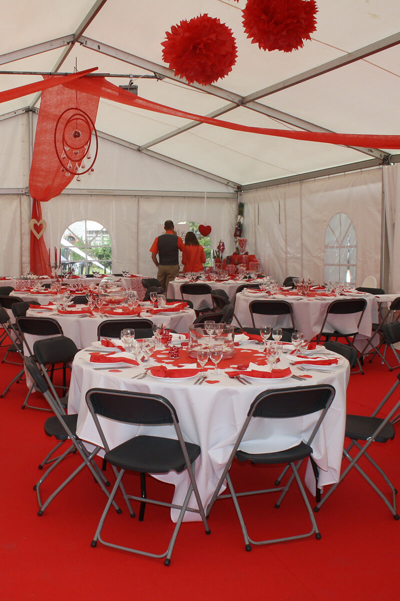 Intérieur d’une tente décorée pour un événement (probablement un mariage ou une réception), avec des tables rondes, des nappes blanches et rouges, et des décorations suspendues au plafond.